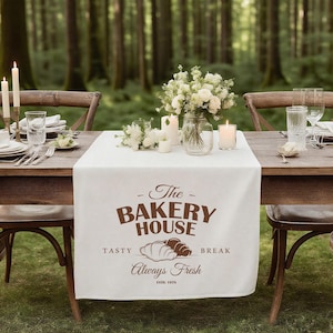 Op de afbeelding: Een witte tafelloper met bruine tekst met de tekst "The Bakery House" en "Always Fresh". De loper ligt op een houten tafel die is gedekt voor een maaltijd, met kaarsen, bloemen en glaswerk. De achtergrond is een bos.