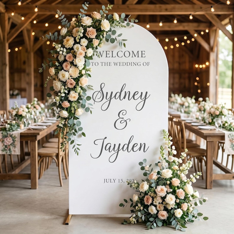 Puede incluir: Un letrero de bienvenida de boda en forma de arco blanco con el texto "WELCOME TO THE WEDDING OF Sydney & Jayden JULY 15, 2024" en letras gris oscuro. El letrero est&aacute; decorado con arreglos de rosas color crema y melocot&oacute;n y hojas de eucalipto verdes.