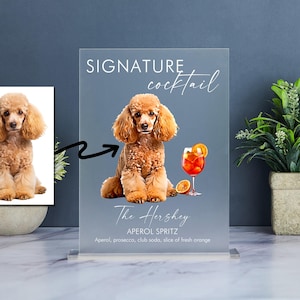 May include: A printable sign with a brown poodle sitting on a gray background. The sign says "SIGNATURE cocktail The Hershey APEROL SPRITZ Aperol, prosecco, club soda, slice of fresh orange".