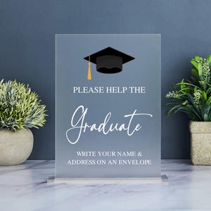 May include: A clear acrylic sign with a black graduation cap graphic and the text "Please help the Graduate. Write your name & address on an envelope."