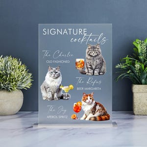 May include: A clear acrylic sign with the text "SIGNATURE cocktails" at the top. Three cocktails are pictured with cats drinking them. The cocktails are "The Charlie" (Old Fashioned), "The Rufus" (Beer Margarita), and "The Joy" (Aperol Spritz).
