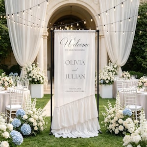 May include: A tall, rectangular white fabric wedding welcome sign suspended on a black metal frame. The sign features black text reading Welcome TO OUR WEDDING OLIVIA and JULIAN JULY 15, 2026 ONEIDA, NEW YORK. The fabric has a ruffled bottom edge.