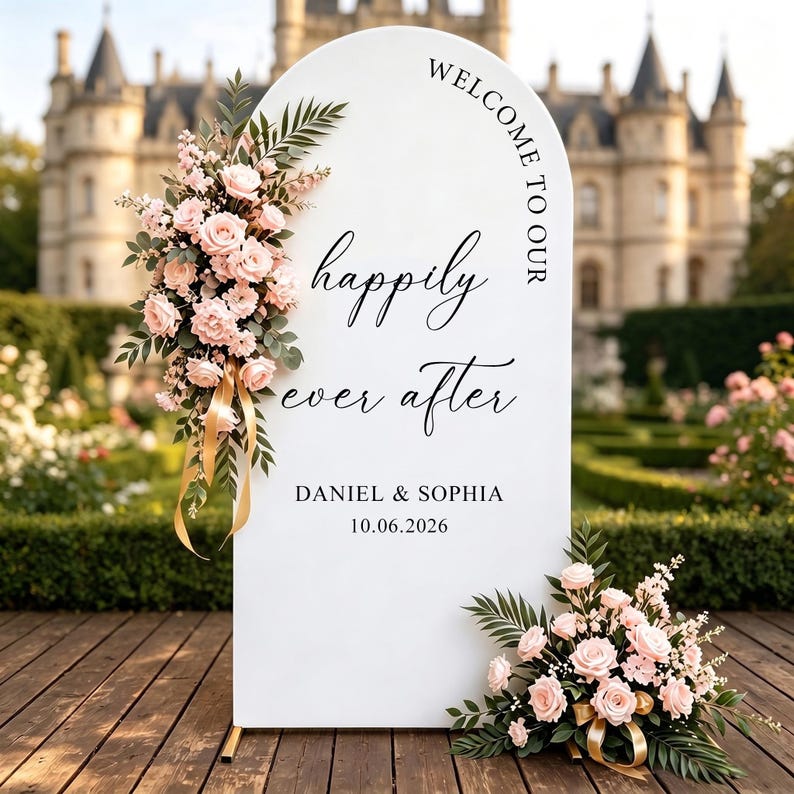 May include: A white arched welcome sign featuring the black text "WELCOME TO OUR happily ever after DANIEL & SOPHIA 10.06.2026". The sign is decorated with two arrangements of light pink roses, green foliage, and gold ribbons on the left and bottom.