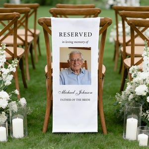 May include: White fabric chair banner with the word "RESERVED" and a photo of a man. The banner also includes the text "in loving memory of", "Michael Johnson", and "FATHER OF THE BRIDE". The banner is draped over a wooden chair.