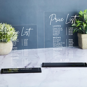 May include: Two clear acrylic signs with black bases displaying a price list for lash and brow services. The signs list prices for classic and volume lash sets, fills, and removal, as well as brow shaping, tinting, microblading, and 3D feathering. The signs also list prices for waxing services, including lip, chin, and eyebrow waxing.