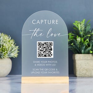 Könnte beinhalten: Ein Schild aus mattiertem Acryl mit Holzsockel. Das Schild hat eine gewölbte Oberseite und trägt die Worte "CAPTURE the love" in eleganter Schrift. Unter dem Text wird ein QR-Code angezeigt, mit den Worten "SHARE YOUR PHOTOS & VIDEOS WITH US! SCAN THE QR CODE & UPLOAD YOUR FAVORITES".