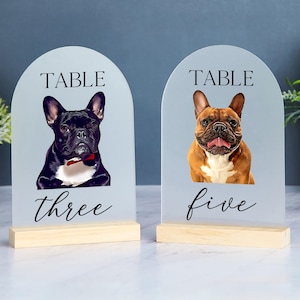May include: Two white acrylic table number signs with a black base. The signs feature a black and white French bulldog and a brown and white French bulldog. The signs read "Table Three" and "Table Five".