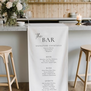 May include: A white bar menu with the text "The BAR" and a list of signature cocktails, beer, wine, and spirits. The bar is set up with stools, a vase of flowers, and a lit candle. The background is a wooden wall.