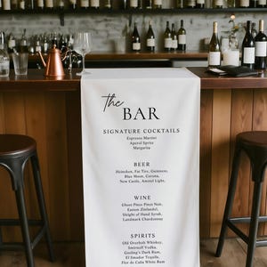 May include: A bar scene with a white cloth displaying a drink menu. The menu lists signature cocktails, beer, wine, and spirits. The bar has a wooden counter, stools, and shelves of bottles.