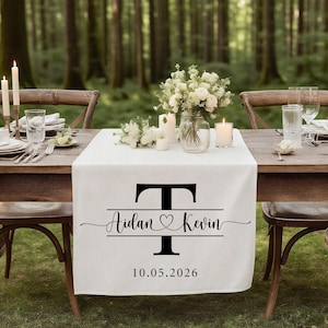 May include: A white table runner featuring a black initial "T", the names "Aidan & Kevin" with a heart symbol, and the date "10.05.2026". The table is set for a wedding with flowers and candles.