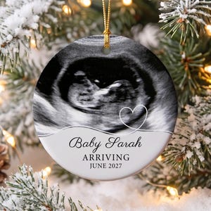 May include: A round ceramic ornament featuring a black and white ultrasound image of a fetus, a white heart outline, and the text Baby Sarah ARRIVING JUNE 2027. The ornament is suspended by a gold cord and has a smooth, glossy finish.