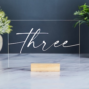 Puede incluir: Un número de mesa de acrílico transparente con la palabra "three" escrita en cursiva blanca. El número está en un pequeño soporte de madera.