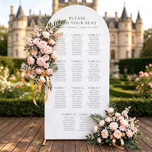 May include: A white arched wedding seating chart sign with black text reading PLEASE FIND YOUR SEAT, Nicole + Michael, August 10, 2026, and a list of tables 1 through 10. The sign is decorated with two floral arrangements of light pink roses and green leaves.