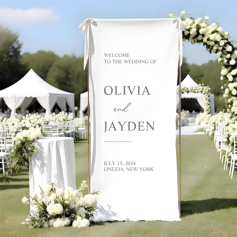 May include: A white wedding welcome sign with the names Olivia and Jayden, set against a backdrop of a wedding ceremony. The sign includes the date July 15, 2024, and the location, Oneida, New York. The sign is framed by a gold metal structure.