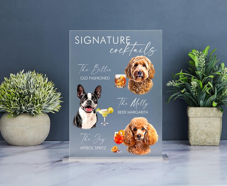 May include: A clear acrylic sign with the text "SIGNATURE cocktails" at the top. The sign features three cocktails with dog illustrations and names: "The Billie Old Fashioned", "The Molly Beer Margarita", and "The Joy Aperol Spritz".