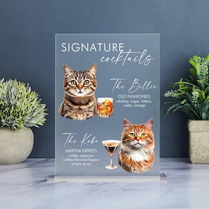 May include: A clear acrylic sign with the text "SIGNATURE cocktails" at the top. The sign features two illustrated cats, one with a martini glass and the other with an old fashioned glass. The text below the cats describes the cocktails, "The Billie" and "The Kobe", with their ingredients.