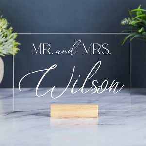 May include: A clear acrylic sign with a wooden stand. The sign says "MR. and MRS. Wilson" in white lettering.
