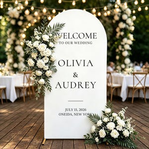 Puede incluir: Un letrero de bienvenida de boda blanco en forma de arco con texto negro que dice WELCOME TO OUR WEDDING, OLIVIA & AUDREY, JULY 15, 2026, ONEIDA, NEW YORK. El letrero est&aacute; decorado con dos arreglos de rosas blancas y follaje verde.