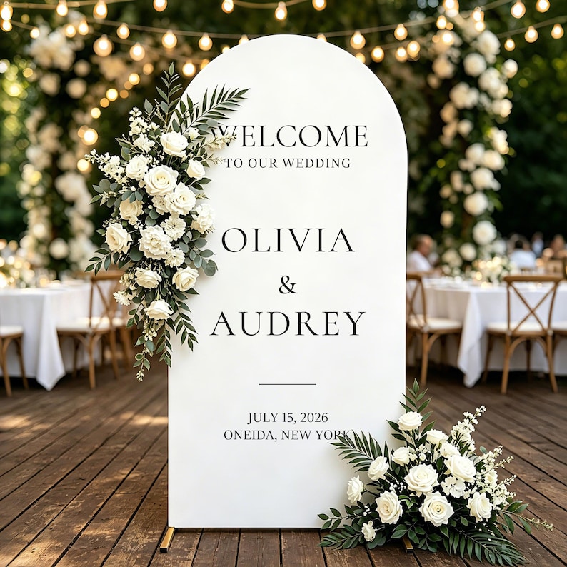 Puede incluir: Un letrero de bienvenida de boda blanco en forma de arco con texto negro que dice WELCOME TO OUR WEDDING, OLIVIA & AUDREY, JULY 15, 2026, ONEIDA, NEW YORK. El letrero est&aacute; decorado con dos arreglos de rosas blancas y follaje verde.