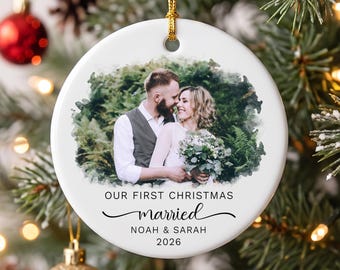 May include: A round white ceramic ornament featuring a watercolor-style photo of a couple in wedding attire. The text reads "OUR FIRST CHRISTMAS married NOAH & SARAH 2026". The ornament has a gold string for hanging and a smooth, glossy finish.
