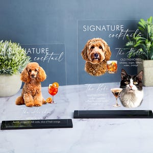 May include: Two clear acrylic signs with black bases, featuring illustrations of a dog and a cat, each with a cocktail recipe. The dog sign says "The Her chay APEROL SPRITZ" and the cat sign says "The Bellie OLD FASHIONED" and "The Kobe MARTINI EXPRESS".