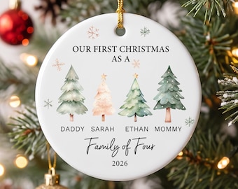 May include: A round white ceramic Christmas ornament featuring a gold hanging string. The text reads OUR FIRST CHRISTMAS AS A, Family of Four, 2026, and names DADDY, SARAH, ETHAN, MOMMY. Four watercolor-style pine trees and small snowflakes are printed.