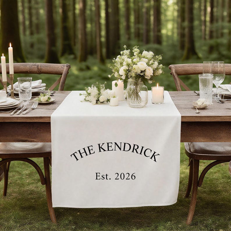 Op de afbeelding: Een witte tafelloper met de zwarte tekst "THE KENDRICK" en "Est. 2026". De tafel is gedekt met witte borden, bestek, kaarsen en bloemstukken. De sc&egrave;ne speelt zich buiten af met een bos op de achtergrond.