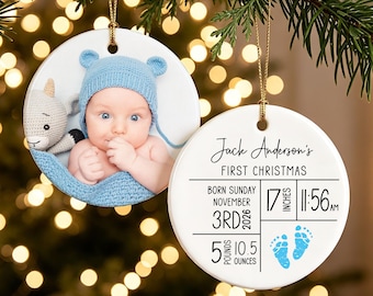 May include: Two round white ceramic ornaments with gold hanging strings. One features a photo of a baby in a blue knit hat and blanket with a stuffed animal. The other displays text: Jack Anderson's FIRST CHRISTMAS, birth details, and blue footprints.