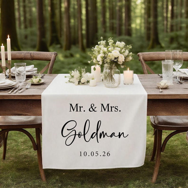 Op de afbeelding: Een witte tafelloper met de tekst "Mr. & Mrs. Goldman 10.05.26" in zwart script. De loper ligt op een houten tafel die is gedekt voor een bruiloftsreceptie, met kaarsen, bloemen en glaswerk. De achtergrond is een vage bos.