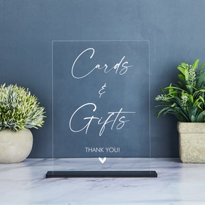 May include: Clear acrylic sign with white text that reads "Cards & Gifts Thank You!" with a small white heart at the bottom. The sign is on a black stand.
