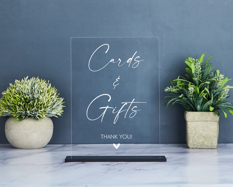 May include: Clear acrylic sign with white text that reads "Cards & Gifts Thank You!" with a small white heart at the bottom. The sign is on a black stand.