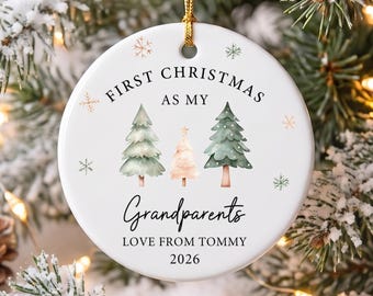 Peut inclure: Une décoration ronde en céramique blanche avec le texte FIRST CHRISTMAS AS MY Grandparents LOVE FROM TOMMY 2026. Le motif présente trois sapins de style aquarelle en vert et rose clair, entourés de petites illustrations de flocons.