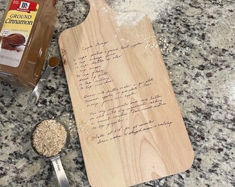 May include: A wooden cutting board with a handwritten recipe for Apple Crisp. The recipe includes ingredients like butter, sugar, flour, and oats. The board is a light brown color and has a handle on the top.