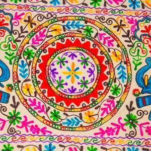 May include: Close-up of a beige textile with vibrant embroidery. The design features concentric circles in red, green, and yellow, adorned with floral and leaf motifs in pink, blue, and purple. The edges have a repeating wave pattern.