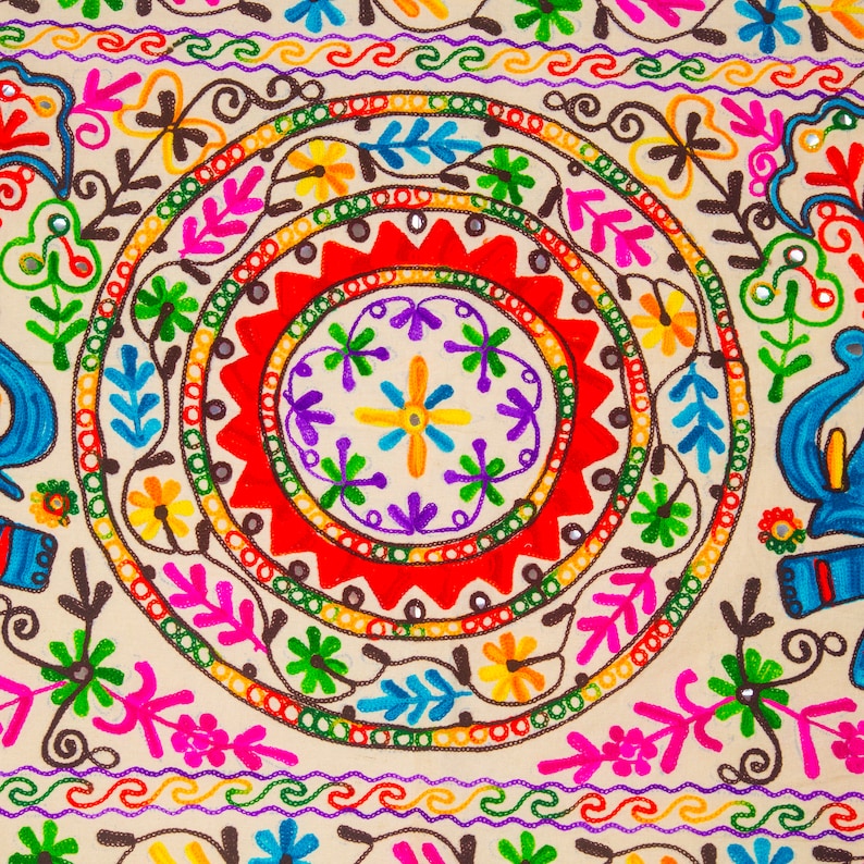 May include: Close-up of a beige textile with vibrant embroidery. The design features concentric circles in red, green, and yellow, adorned with floral and leaf motifs in pink, blue, and purple. The edges have a repeating wave pattern.