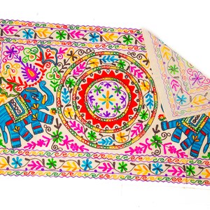 May include: Embroidered rectangular textile with a cream-colored background. The design features a central circular pattern with red, yellow, and purple accents. Two blue elephants are on either side. The border is decorated with colorful floral and leaf motifs.