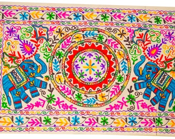 May include: Embroidered rectangular textile featuring a central circular design and two blue elephants. The textile is adorned with vibrant colors including red, yellow, green, and pink, with intricate floral and geometric patterns. The background is a light beige.