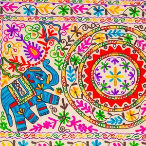 May include: Close-up of a colorful, embroidered textile featuring a blue elephant and intricate floral patterns. The design includes vibrant hues of red, yellow, green, and purple, with a cream-colored background. The textile has a circular design in the center.