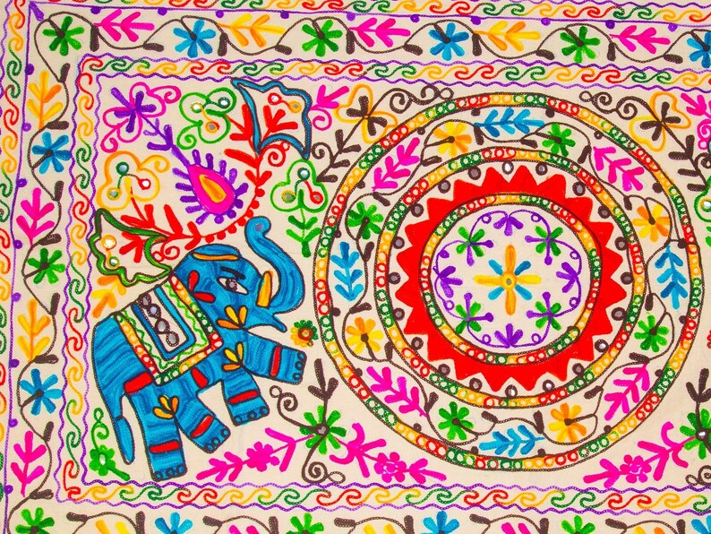 May include: Close-up of a colorful, embroidered textile featuring a blue elephant and intricate floral patterns. The design includes vibrant hues of red, yellow, green, and purple, with a cream-colored background. The textile has a circular design in the center.