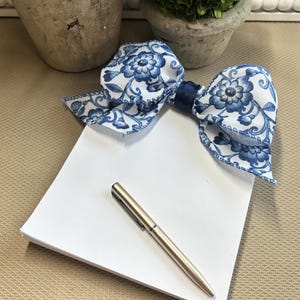 May include: A white notepad with a blue and white floral bow on top and a silver pen lying on the notepad.