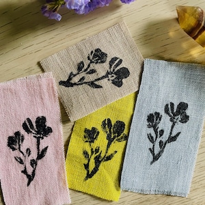 Könnte beinhalten: Vier Stoffetiketten in Rosa, Beige, Gelb und Blau-Grau, jeweils mit einem schwarzen Blumendesign. Die Etiketten sind auf einer hellen Holzoberfläche angeordnet, mit lila Blumen und einem gelben Kristall im Hintergrund. Die Etiketten sind rechteckig.