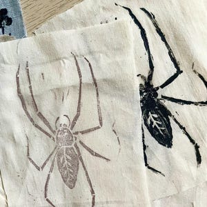 May include: Close-up of fabric squares with spider prints in brown and black ink. The spiders are depicted with detailed legs and body patterns. The fabric has a natural, off-white color, and the prints have a handmade, artistic quality.
