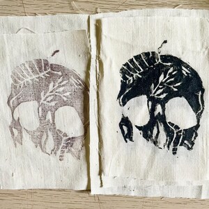 Può includere: Due quadrati di tessuto affiancati, ciascuno con una stampa di teschio. Uno è viola tenue, l'altro nero. Il disegno del teschio include dettagli di foglie e steli. Il tessuto è di un colore bianco sporco naturale, con una trama visibile.