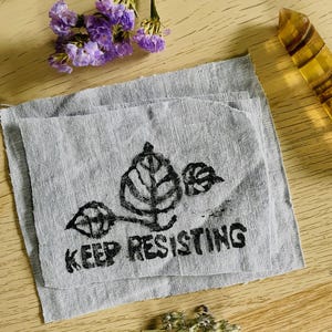 May include: Two gray fabric squares with a black leaf design and the words "KEEP RESISTING" printed on them. The image also includes purple flowers, a yellow crystal, and small dried plants, all arranged on a wooden surface.