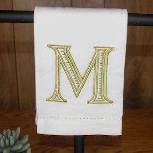 May include: A white hand towel featuring a large, centred, embroidered letter M in a metallic gold thread. The letter has a decorative, lined interior pattern. The towel is draped over a black horizontal bar and has a simple stitched hem at the bottom.