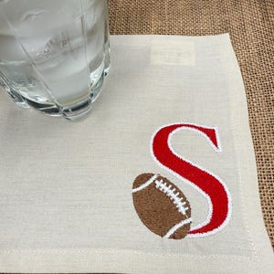 May include: A cream linen coaster featuring an embroidered American football and the letter "S" in red. A clear glass with ice rests on the coaster, which is on a burlap surface.