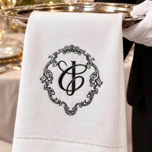 May include: A white linen napkin featuring a black embroidered monogram of the letters C and J, enclosed within an ornate, decorative black circular frame. The napkin has a hemstitched border detail along the bottom edge.