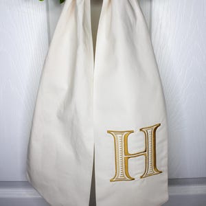 May include: An off-white fabric door hanger featuring a large, embroidered gold letter H with a decorative border. The fabric is gathered at the top and hangs vertically, displaying a smooth, matte finish on the textile surface.