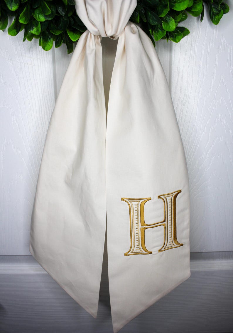 May include: An off-white fabric door hanger featuring a large, embroidered gold letter H with a decorative border. The fabric is gathered at the top and hangs vertically, displaying a smooth, matte finish on the textile surface.