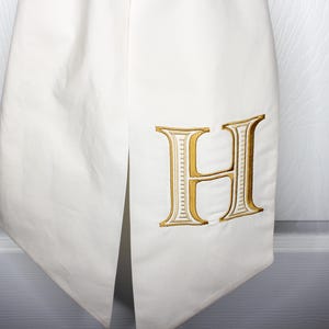 May include: A white fabric item featuring a large, embroidered letter H in a metallic gold thread with white detailing. The fabric has a smooth texture and a vertical slit, showcasing a clean, structured design with precise stitching on the letter.